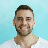 Smiling man with short brown hair and beard wearing a white shirt against a light blue background.